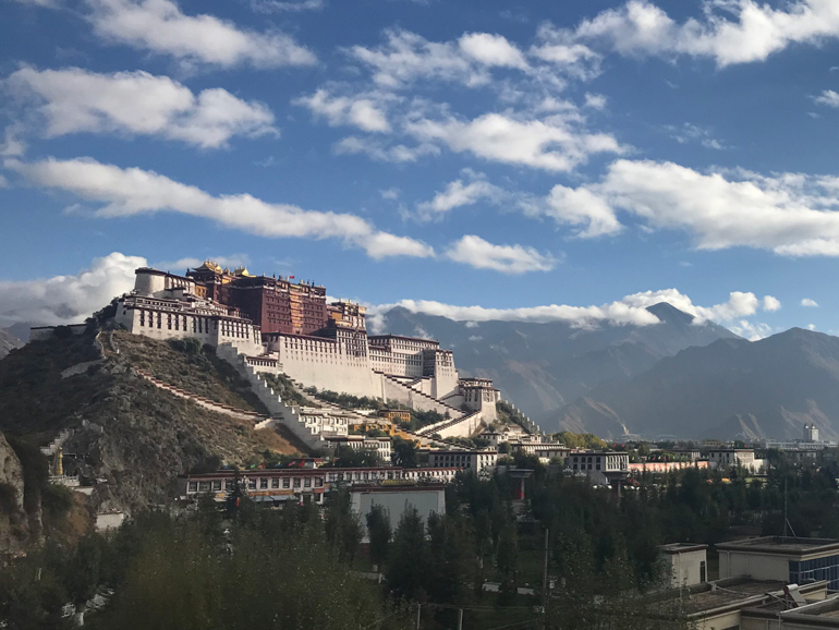 Tibets kulturelles Erbe: Der Potala-Palast inmitten der Berge Blick auf den Potala-Palast in Lhasa, umgeben von modernen Gebäuden und Bergen im Hintergrund, bei teilweise bewölktem Himmel.