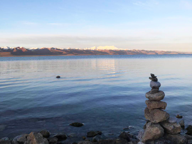 Heilige Gewässer Tibets: Ein Spiegelbild der Göttlichkeit Ein ruhiger tibetischer See, umgeben von schneebedeckten Bergen, mit einem spirituellen Steinhaufen im Vordergrund.