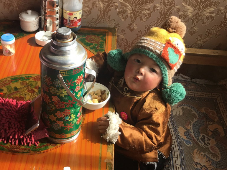Kulturelle Zärtlichkeit: Ein tibetisches Kind im Alltag Tibetisches Kind in einem bunten Hut und einer braunen Jacke isst Brot, ein Bild, das die kulturelle Wertschätzung von Kindern widerspiegelt.