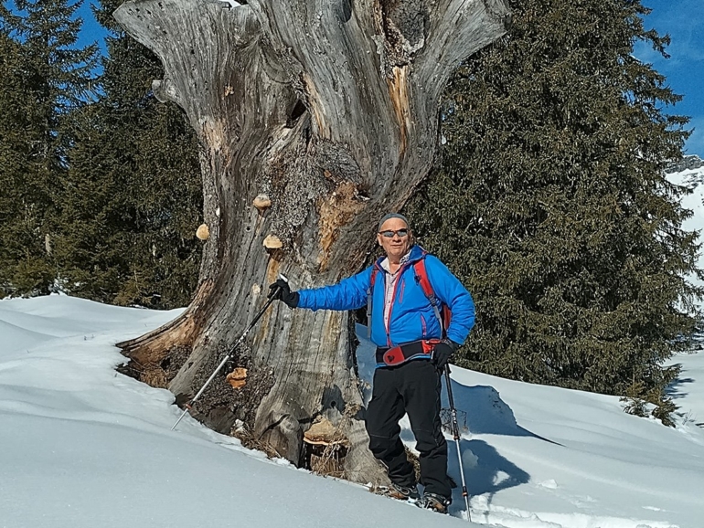 Armin posiert bei einer Schneeschuhwanderung in Damüls neben einem alten Baumstumpf im Schnee.